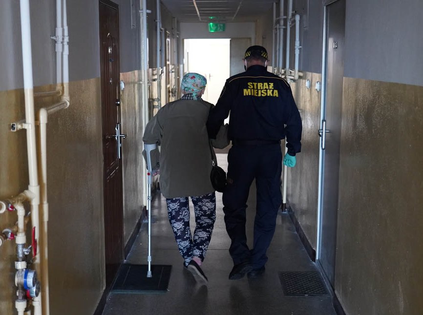 Strażnik miejski prowadzi korytarzem budynku starszą kobietę z laską. Zdjęcie oryginalne.