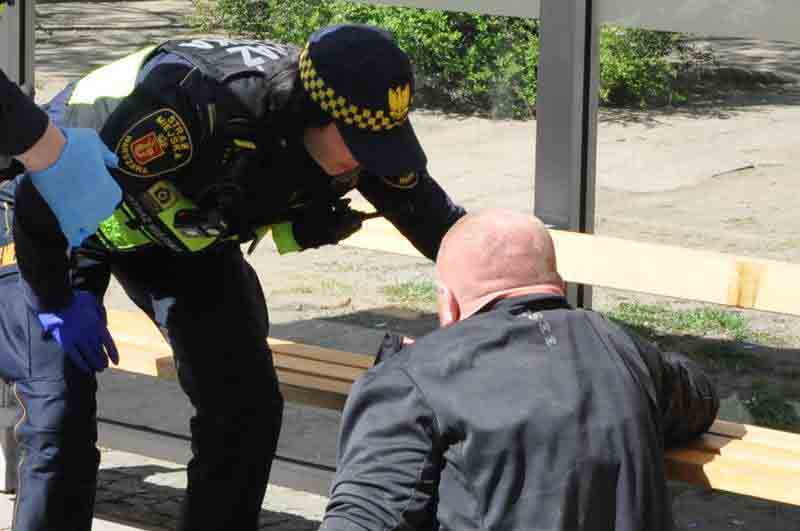 Zdjęcie ilustracyjne: strażniczka miejska pochyla się nad mężczyzną siedzącym na ziemi przy przystanku.