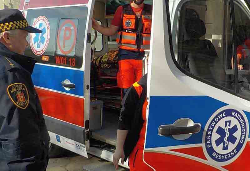 Zdjęcie ilustracyjne: strażnik miejski stojący przy uchylonych drzwiach karetki, w których stoi ratownik pogotowia ratunkowego.