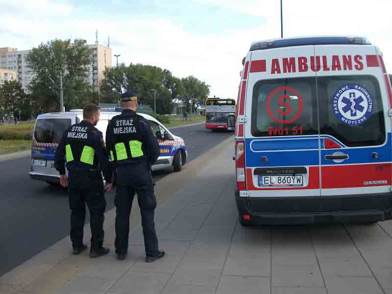 Zdjęcie ilustreacyjne- dwaj strażnicy stojący przy karetce pogotowia ratunkowego. W tle radiowóz straży miejskiej oraz autobus miejski.
