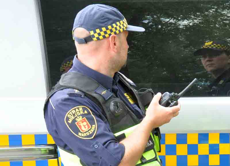 Strażnik miejski z radiotelefonem w ręku na tle radiowozu straży miejskiej- zdjęcie ilustracyjne