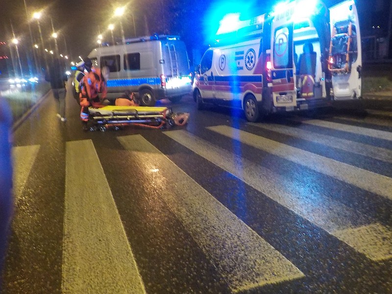 Zdjęcie ilustracyjne: noc, przejście dla pieszych. W tle ratownicy medyczni przy złożonych noszach, karetka pogotowia z włączonymi światłami sygnalizacyjnymi oraz policyjny radiowóz.