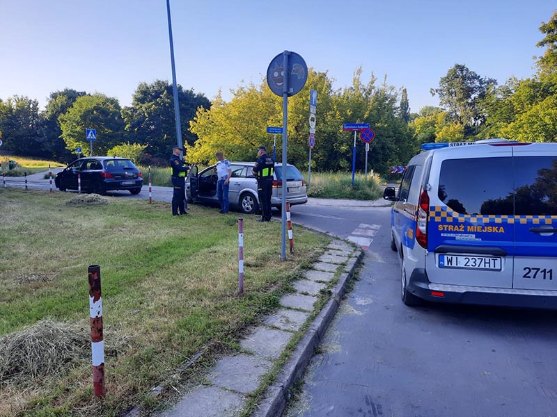 Ujęty mężczyzna stoi przy swoim samochodzie, obok stoją dwaj strażnicy miejscy. Z prawej strony radiowóz straży miejskiej.