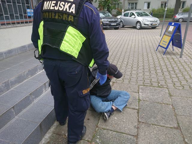 Zdjęcie ilustracyjne: strażnik miejski podchodzący do siedzącego na chodniku przy betonowych schodach mężczyzny.