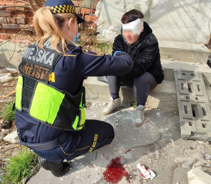 Zdjęcie z interwencji: strażniczka miejska opatruje siedzącą na ziemi kobietę, która ma już na głowie bandaż.