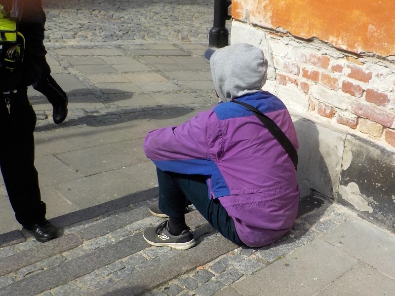 Zdjęcie z interwencji: mężczyzna w fioletowo-niebieskiej kurtce, z szarym kapturem na głowie, siedzi na chodniku przy murze. Obok stoi strażnik miejski.