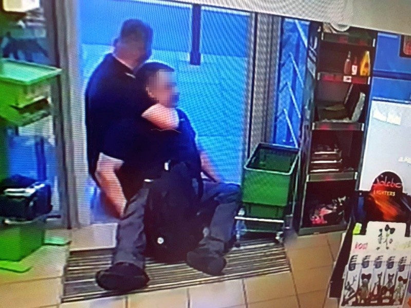 Ujęcie z kamery monitoringu: agresywny mężczyzna siedzi w drzwiach sklepu unieruchomiony chwytem obezwładniającym przez strażnika miejskiego w cywilu.