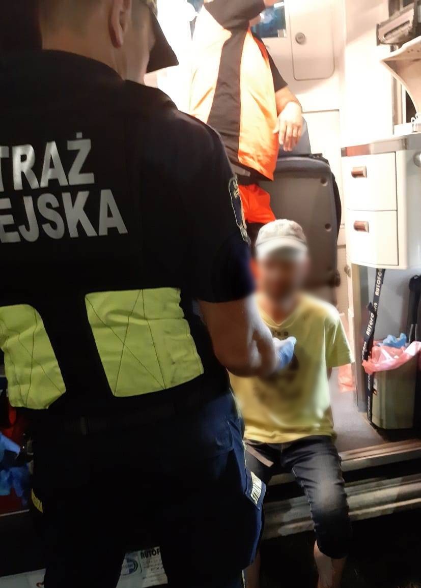 Mężczyzna w żółtym T-shircie siedzi na wejściu do ambulansu, przed którym stoi strażnik miejski (widać plecy). We wnętrzu karetki ratownik pogotowia sięga po coś.