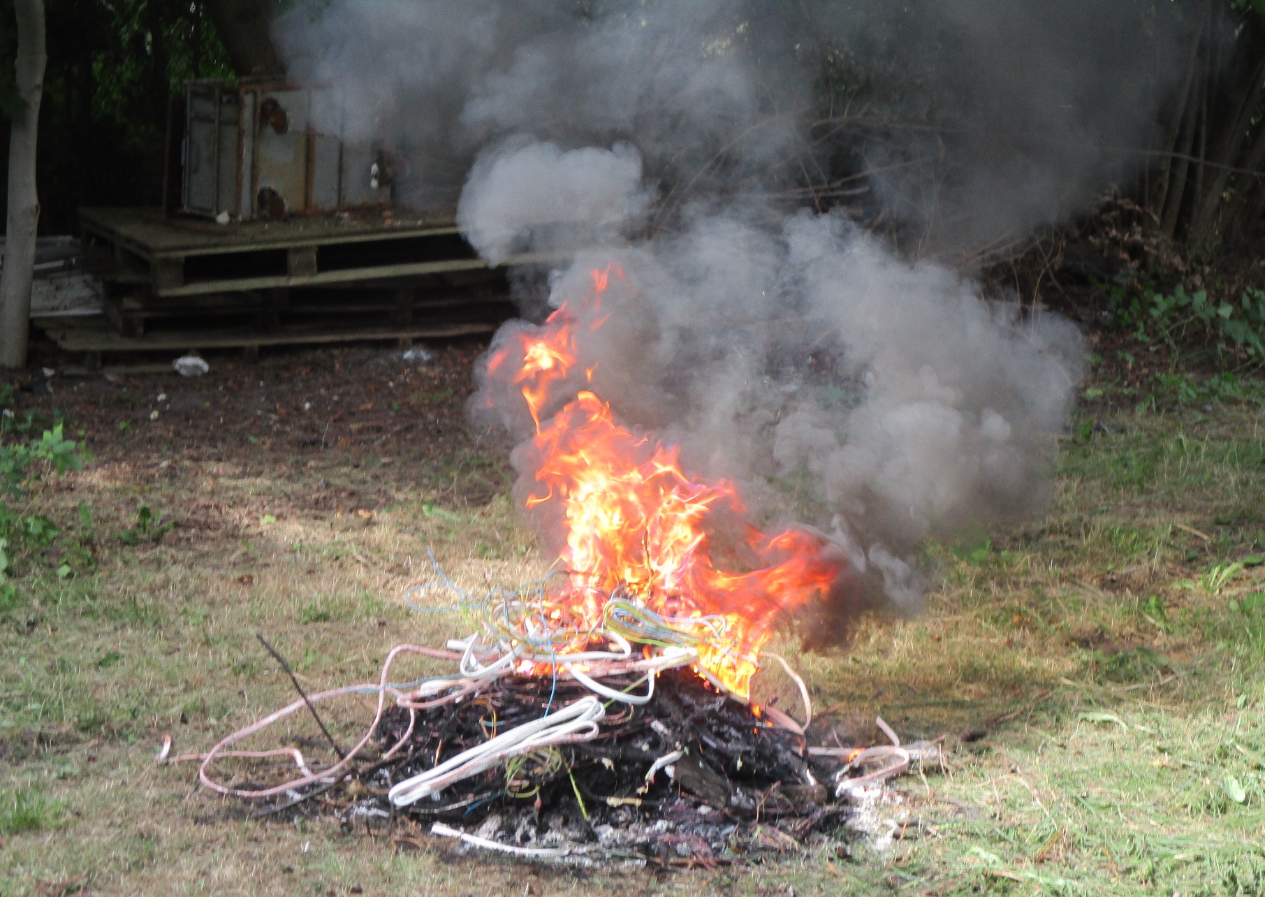 Płonące ognisko, a nad nim ciężki, szary dym. Z ognia wystają kable w plastikowej izolacji.
