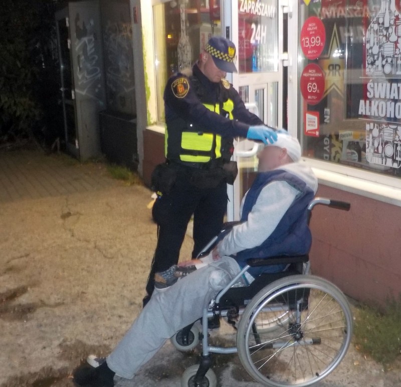 Strażnik miejski opatrujący głowę mężczyzny siedzącego na wózku inwalidzkim.
