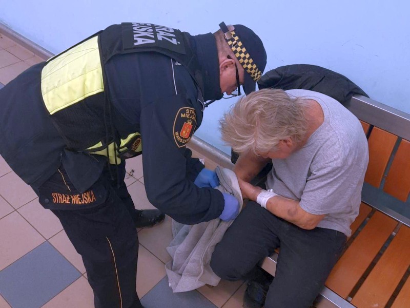 Strażnik pomagający się ubrać siedzącemu na ławce na korytarzu mężczyźnie.