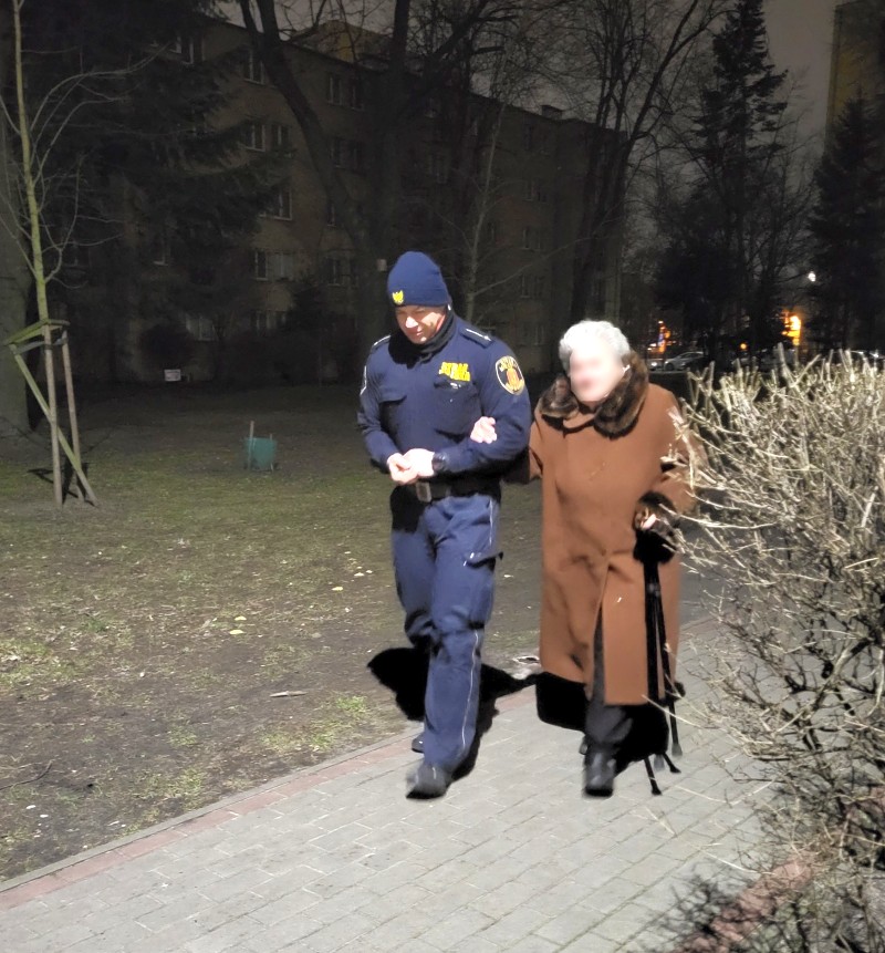 Kobieta w brązowym płaszczu idzie pod rękę ze strażnikiem miejskim.