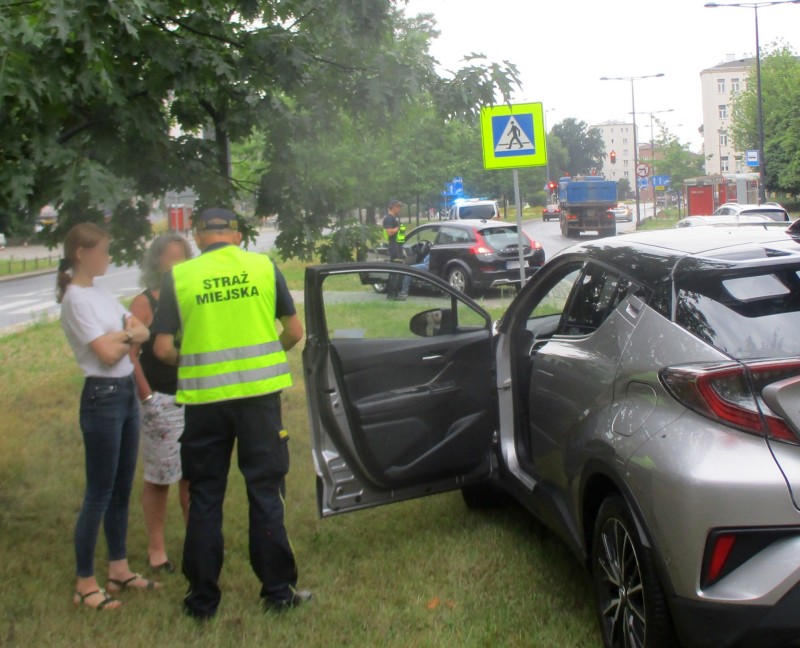 Strażnik miejski obok kobiet uczestniczących w kolizji, stojących przy uszkodzonym samochodzie.