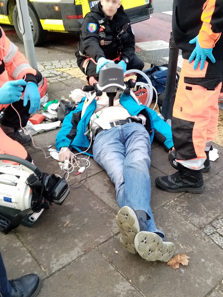 Mężczyzna z aparaturą resuscytacyjną  leży na chodniku wśród ratowników medycznych.