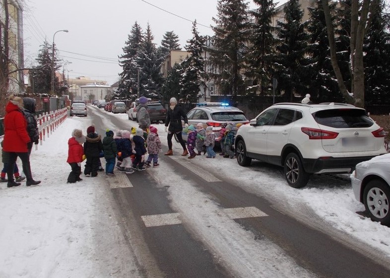 Suw zaparkowany na pasach dla pieszych zmusza wycieczkę dzieci do przeciskania się między samochodami. W drugim planie zaparkowany radiowóz interweniujących strażników miejskich.