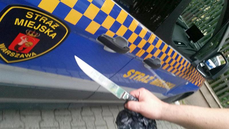Ręka trzymająca nóż na tle radiowozu straży miejskiej- zdjęcie ilustracyjne.