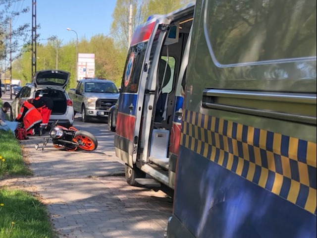 Radiowóz straży miejskiej, w tle karetka pogotowia i leżący na ziemi motocykl i ratownicy udzielający pomocy motocykliście