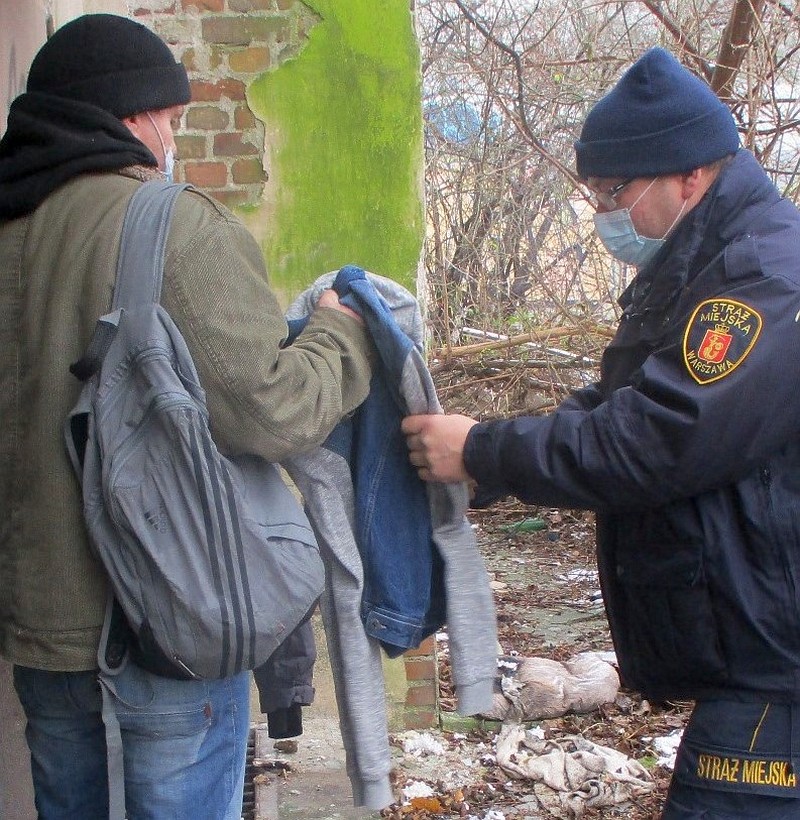 Zdjęcie ilustracyjne: stojący po prawej stronie kadru strażnik miejski podaje bezdomnemu, stojącemu z lewej, bluzę. W tle opuszczony budynek.