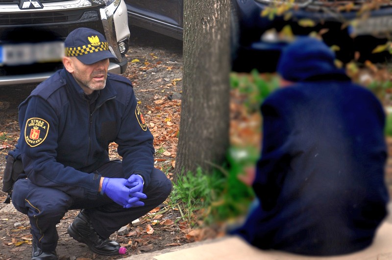 Zdjęcie ilustracyjne: strażnik miejski kuca przy siedzącym na murku mężczyźnie ubranym w granatową bluzę i czapkę.