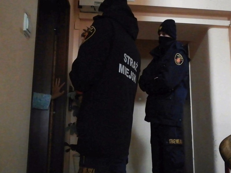 Zdjęcie ilustracyjne: strażnicy miejscy na klatce schodowej budynku, stoją przed drzwiami, z których wychyla się kobieca ręka w szlafroku.
