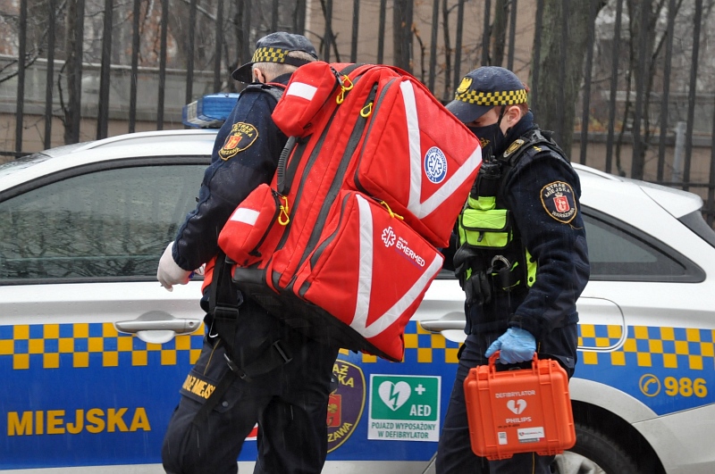 Zdjęcie ilustracyjne: dwóch strażników-ratowników na tle radiowozu straży miejskiej. Jeden trzyma na ramieniu plecak medyczny, drugi w ręku niesie podręczną apteczkę.