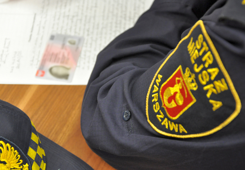 Zdjęcie ilustracyjne: strażnik miejski spisujący notatkę służbową. Na stole, obok ramienia strażnika, leży kobiecy dowód osobisty.