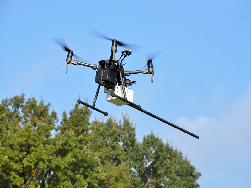 Zdjęcie ilustracyjne: dron z wyposażeniem służącym do wykrywania zanieczyszczeń w dymie unosi się w powietrzu. W tle widoczne zielone korony drzew.