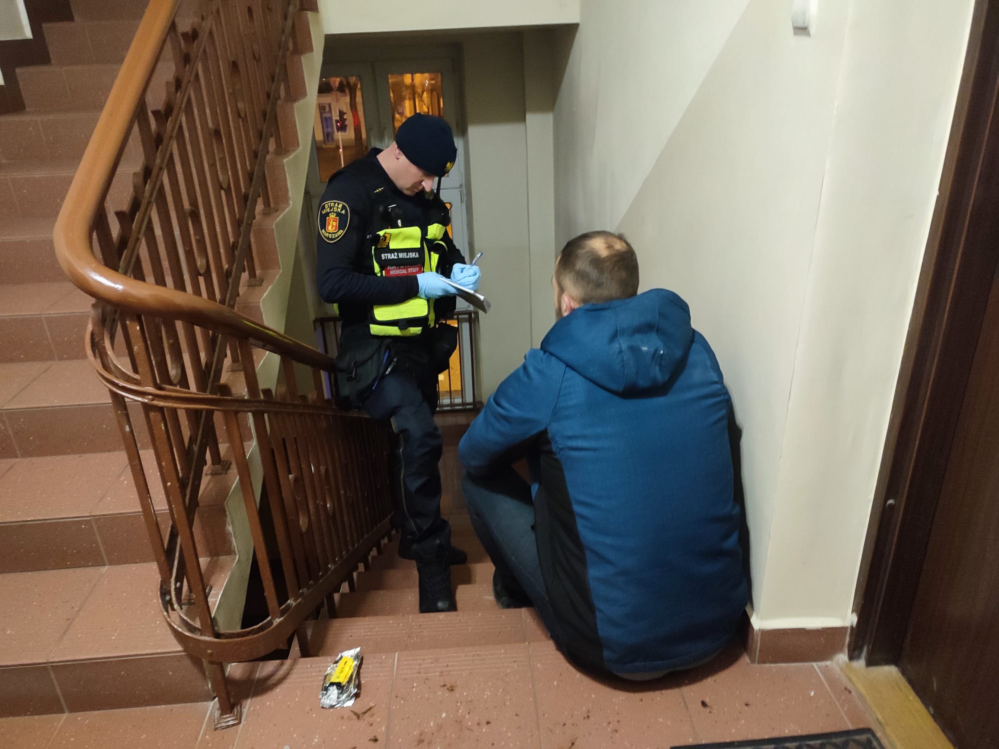 Strażnik miejski spisuje personalia mężczyzny siedzącego na klatce schodowej.