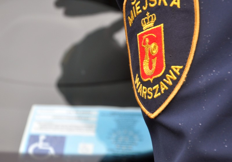 Detal: rękaw z tarczą herbową munduru strażnika miejskiego oglądającego kartę parkingową za szybą pojazdu.