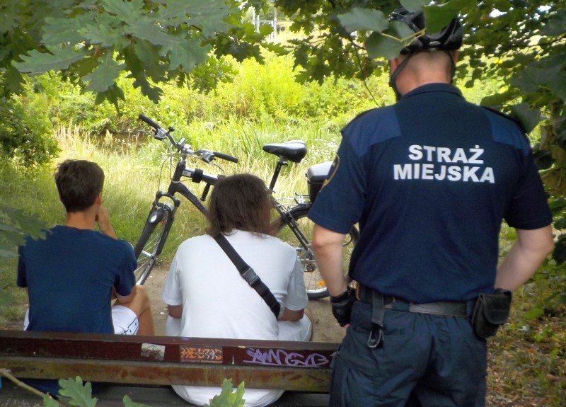 Strażnik miejski stoi za ławką, na której siedzi dwóch chłopców. W tle stoi rower.