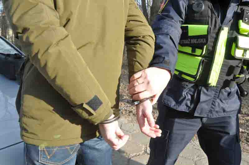 Zdjęcie ilustracyjne: strażnik miejski trzyma za rękę mężczyznę zakutego w kajdanki.