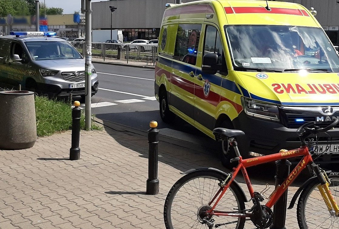 Rower oparty o słup sygnalizacji świetlnej przy przejściu dla pieszych. Obok ambulans pogotowia i radiowóz straży miejskiej.