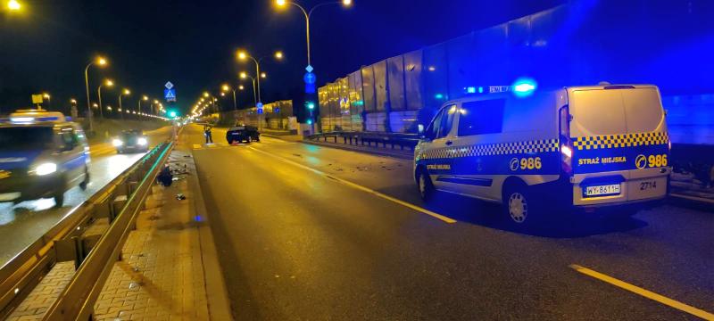 Widok na ulicę Wał Miedzeszyński nocą. Po prawej stronie radiowóz straży miejskiej z włączonymi sygnałami świetlnymi. W tle rozbite ciemne BMW, obok stoi strażnik miejski zabezpieczający miejsce zdarzenia.