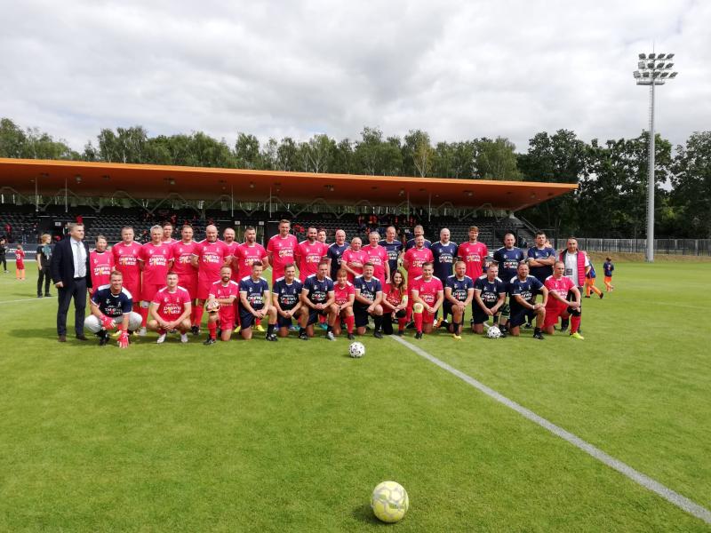 Dwie drużyny piłkarskie pozują do zdjęcia przed meczem- w czerwonych strojach drużyna samorządowców, w granatowych drużyna piłkarzy Hutnika, Legii i Polonii.