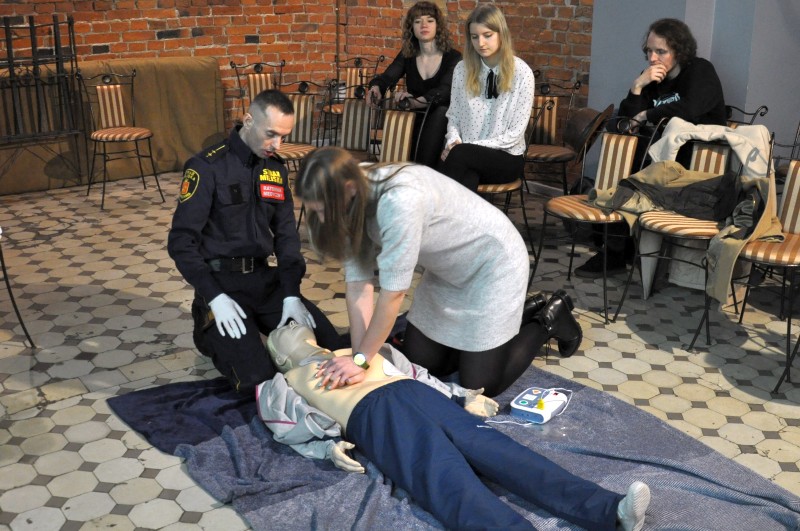 Zdjęcie ze szkolenia: nad fantomem medycznym klęczy młoda kobieta, wykonując masaż serca. Obok niej klęczy ratownik ze Straży Miejskiej. W tle, na krzesłach, obserwujący szkolenie pracownicy teatru "Kamienica".