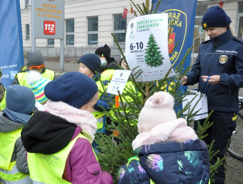 Zdjęcie z rozpoczęcia akcji pod siedzibą Straży Miejskiej przy Młynarskiej: grupa dzieci dekoruje drzewko znaczkami odblaskowymi. Z prawej strony kadru strażniczka miejska z białą torbą papierową podaje dzieciom odblaski.