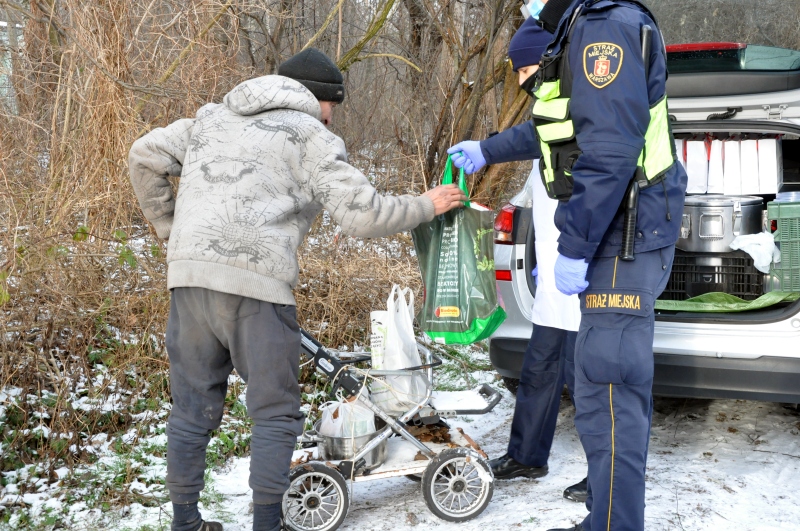 Zdjęcie z akcji: bezdomny w szarej kurtce, z wózkiem dziecięcym służącym mu do transportu rzeczy osobistych, odbiera z rąk strażników paczkę z darami.