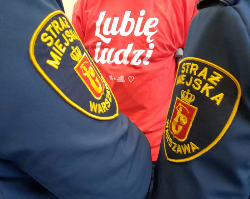 Zdjęcie z akcji: strażnicy w mundurach, w tle czerwona koszulka wolontariusza z białym napisem "Lubię ludzi".