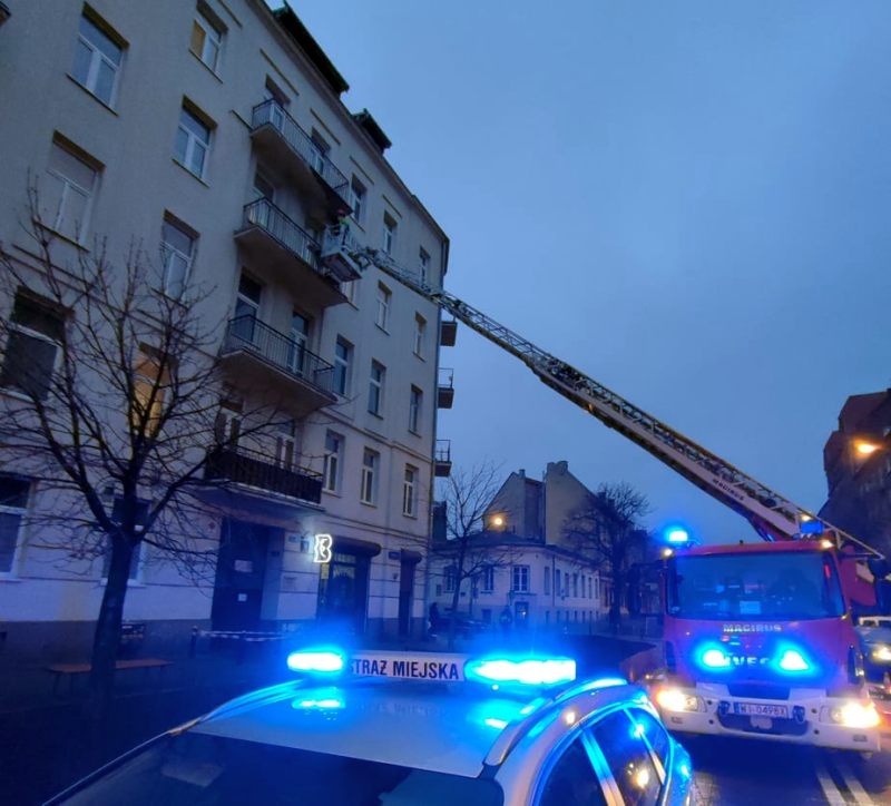 Zdjęcie z interwencji: na pierwszym planie radiowóz straży miejskiej z włączonymi światłami sygnalizacyjnymi, za nim wóz straży pożarnej z rozłożoną drabiną przystawianą do budynku z uszkodzonym balkonem.