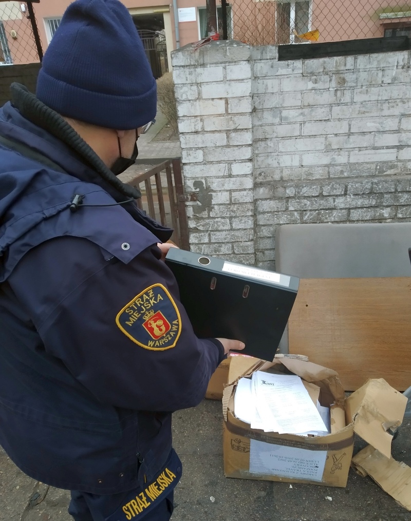 Zdjęcie z interwencji: strażnik miejski stojący przy altanie śmietnikowej, w ręce trzyma czarny skoroszyt z odnalezionymi dokumentami.
