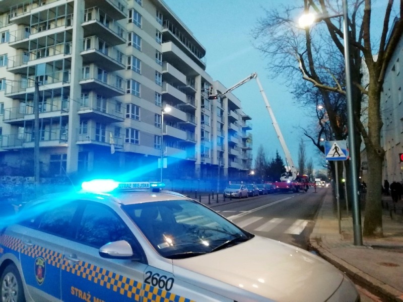 Zdjęcie z interwencji: radiowóz straży miejskiej stojący przy wlocie uliczki prowadzącej do bloku, na balkonie którego wisiała uszkodzona rura.