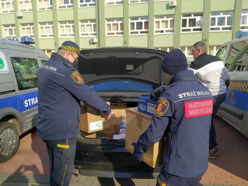 Zdjęcia z przygotowań do wyjazdu: strażnicy miejscy (jeden w cywilnym ubraniu, dwóch w mundurach) pakują paczki z lekarstwami do bagażnika samochodu.