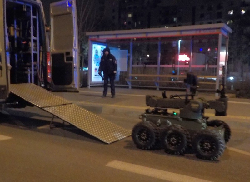 Zdjęcie z interwencji: robot pirotechniczny zjeżdżający z rampy z pojazdu saperów. W tle strażnik miejski i przystanek autobusowy.