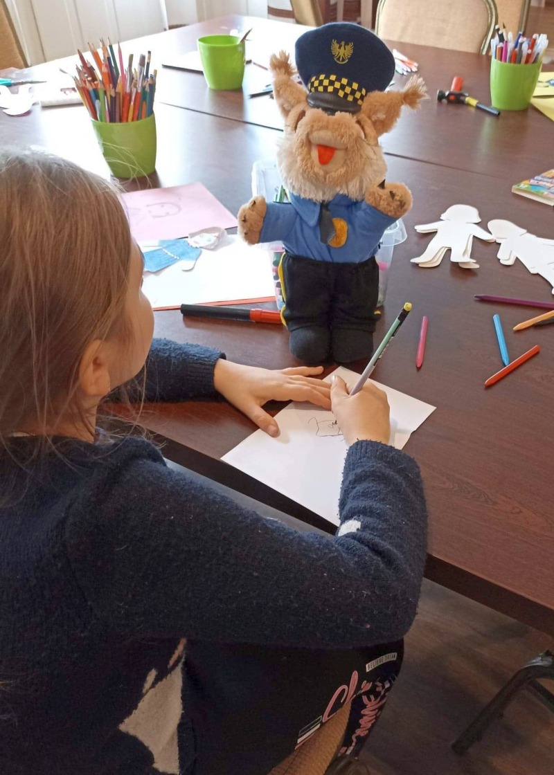 Zdjęcie z zajęć prowadzonych przez Referat Profilaktyki: dziewczynka przy biurku rysuje na kartce, obok niej stoi ryś, maskotka Straży Miejskiej.