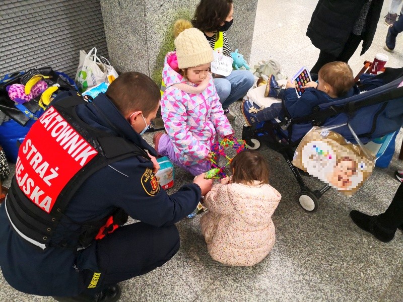 Zdjęcie z akcji pomocowej: w punkcie informacyjnym strażnik miejski kuca obok trójki dzieci, które z matką przyjechały z Ukrainy.