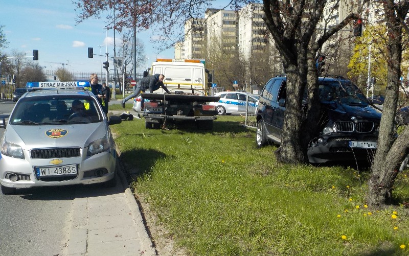 Zdjęcie z interwencji: z prawej strony czarne BMW zatrzymane na dwóch drzewkach, z lewej- stojący na jezdni radiowóz straży miejskiej. Między nimi laweta pomocy drogowej.