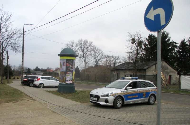 Zdjęcie z interwencji: zwisający kabel leży na chodniku, za nim stoi radiowóz straży miejskiej, w tle zaparkowane samochody i słup ogłoszeniowy.