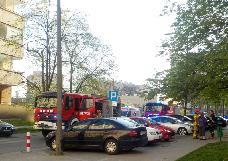 Zdjęcie z interwencji: samochody zaparkowane przy bloku, w którym wybuchł pożar, obok wóz ratowniczy straży pożarnej. Na chodniku strażacy podczas akcji ratowniczej.