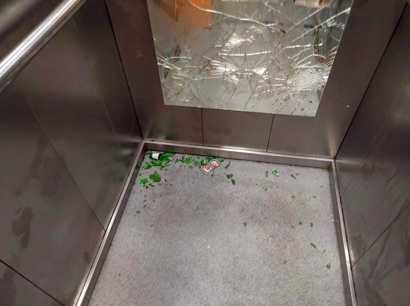 Zdewastowana winda: zbite lustro na ścianie kabiny, rozbita butelka po piwie na podłodze.