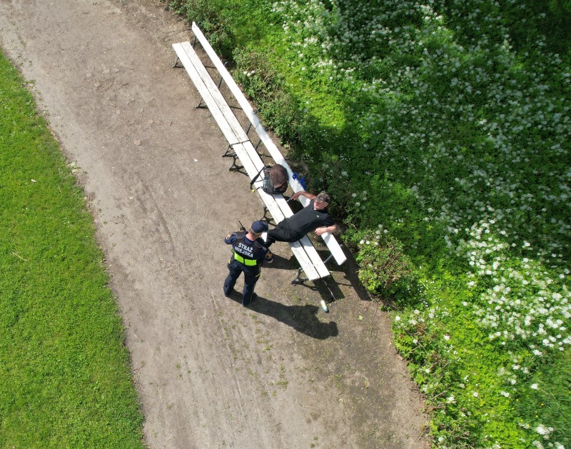 Widok z drona: strażnik miejski podchodzi do siedzącego na ławce w parku mężczyzny.